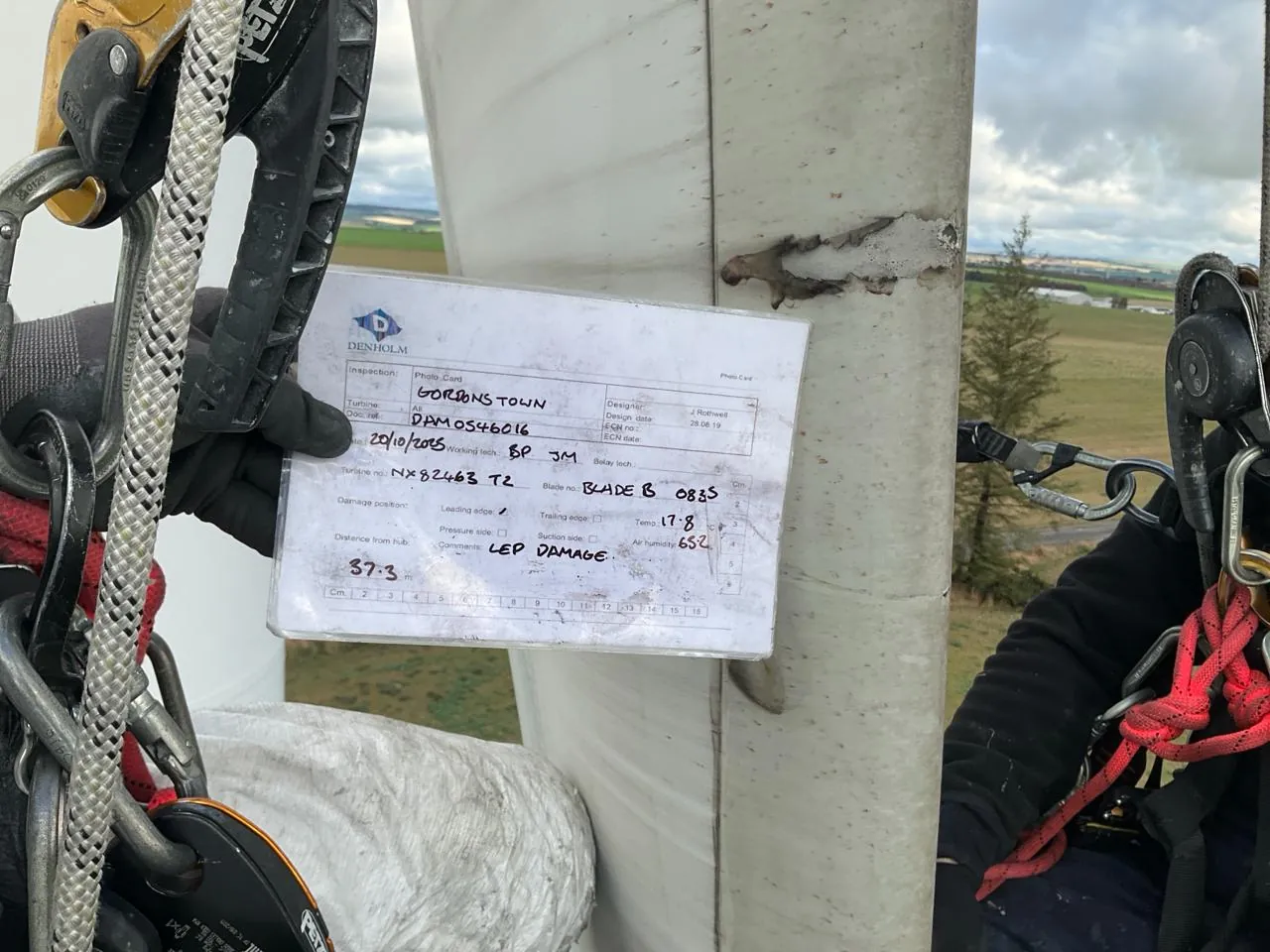 Ein Techniker hält ein ausgefülltes Papier-Inspektionsboard gegen ein Windkraftanlagenblatt in der Höhe — alle Inspektionsfelder sind handschriftlich ausgefüllt, aber ohne BLADE™ verbleiben die Daten als Bild statt als strukturierte Einträge im System