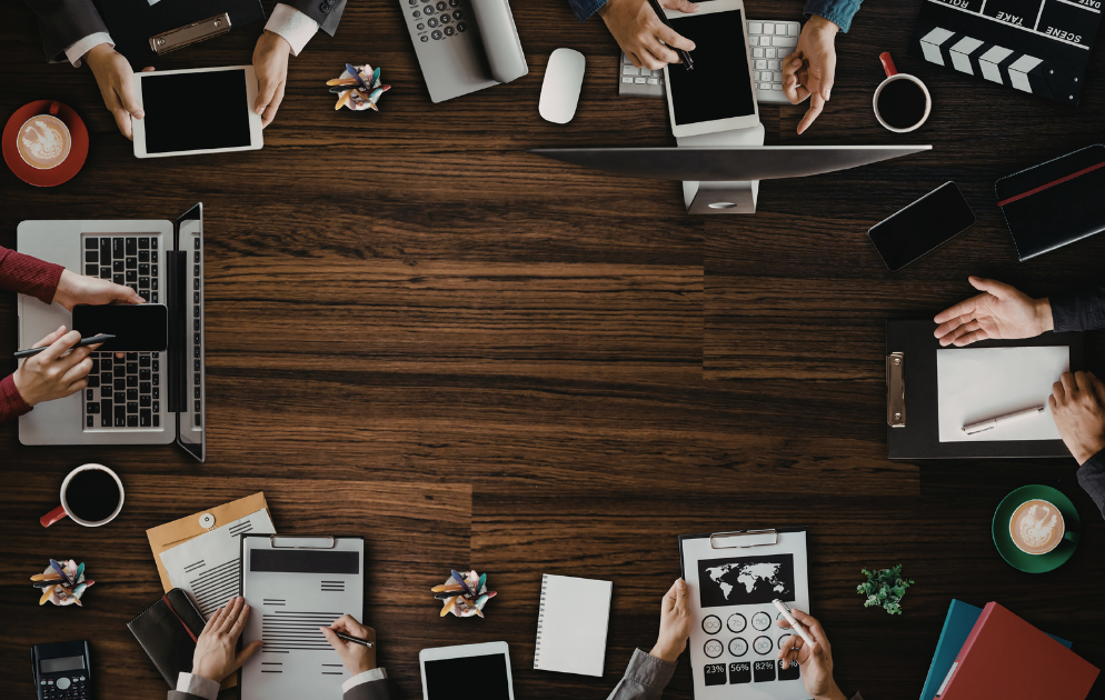 Draufsicht auf ein Team, das an einem Tisch mit Laptops, Tablets, Dokumenten und Kaffee zusammenarbeitet